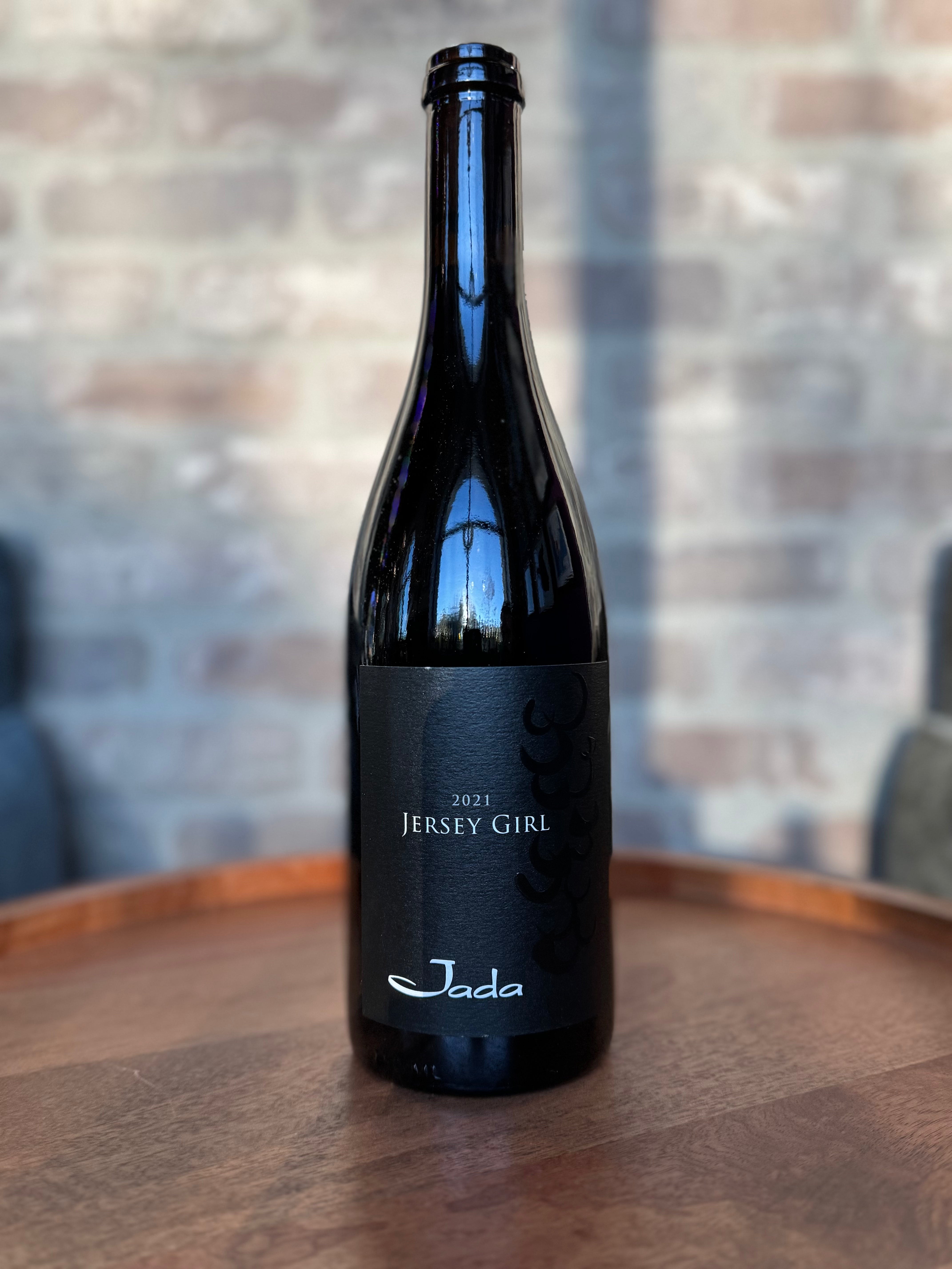 Black wine bottle labeled '2021 Jersey Girl' by Jada on a wooden surface with a blurred brick wall background.