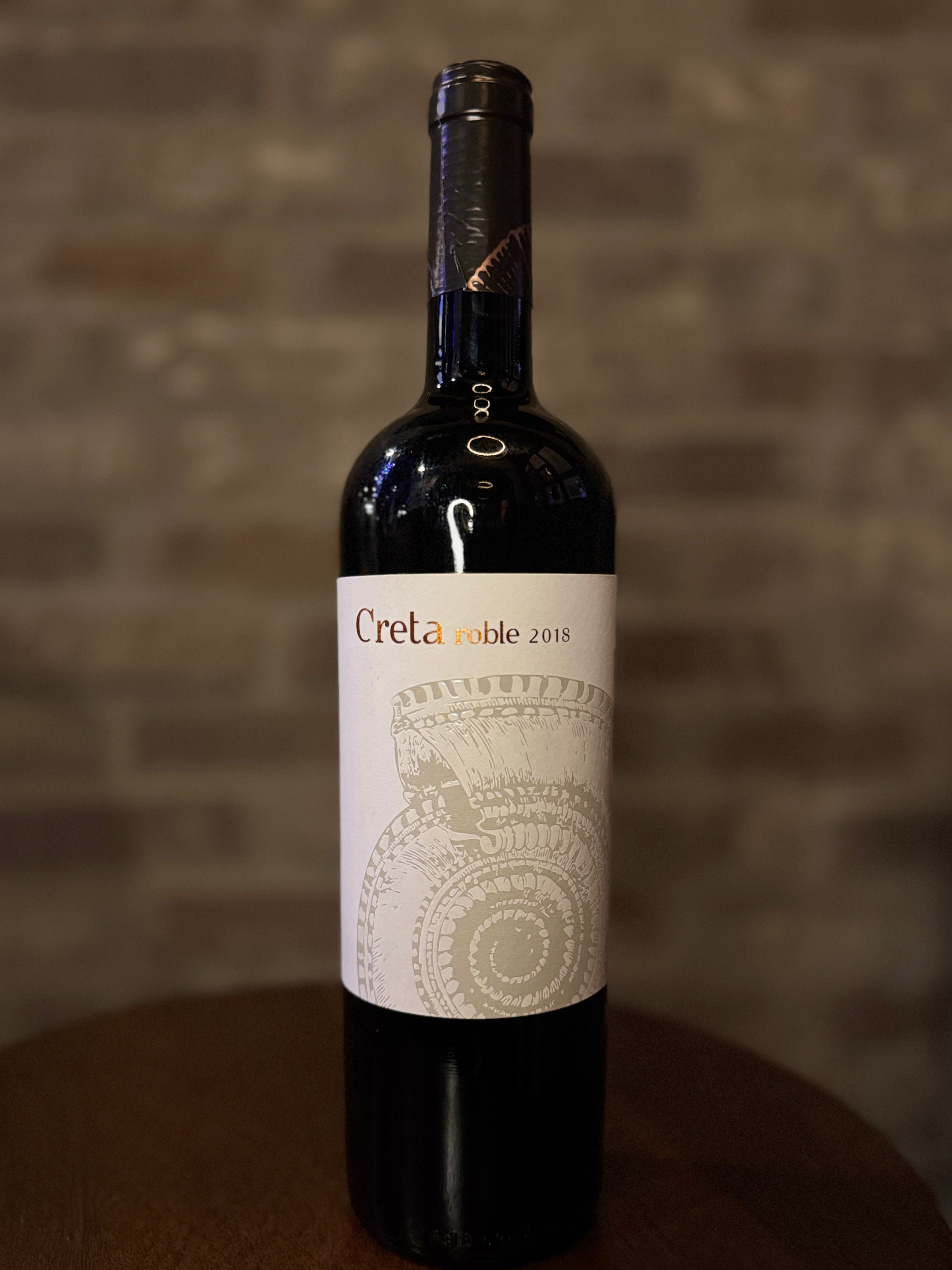 Wine bottle labeled 'Creta Noble 2018' on a wooden surface with a blurred brick wall background.