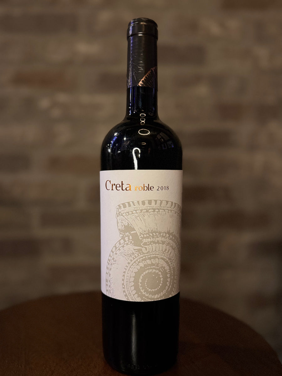 Wine bottle labeled 'Creta Noble 2018' on a wooden surface with a blurred brick wall background.