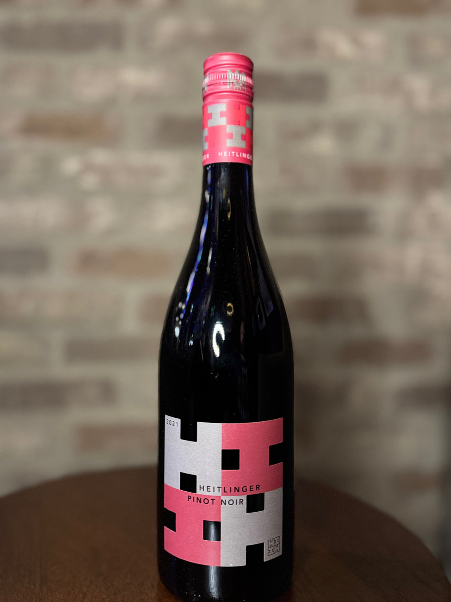Bottle of Heitzinger Pinot Noir wine with a red label on a wooden surface and brick wall background.